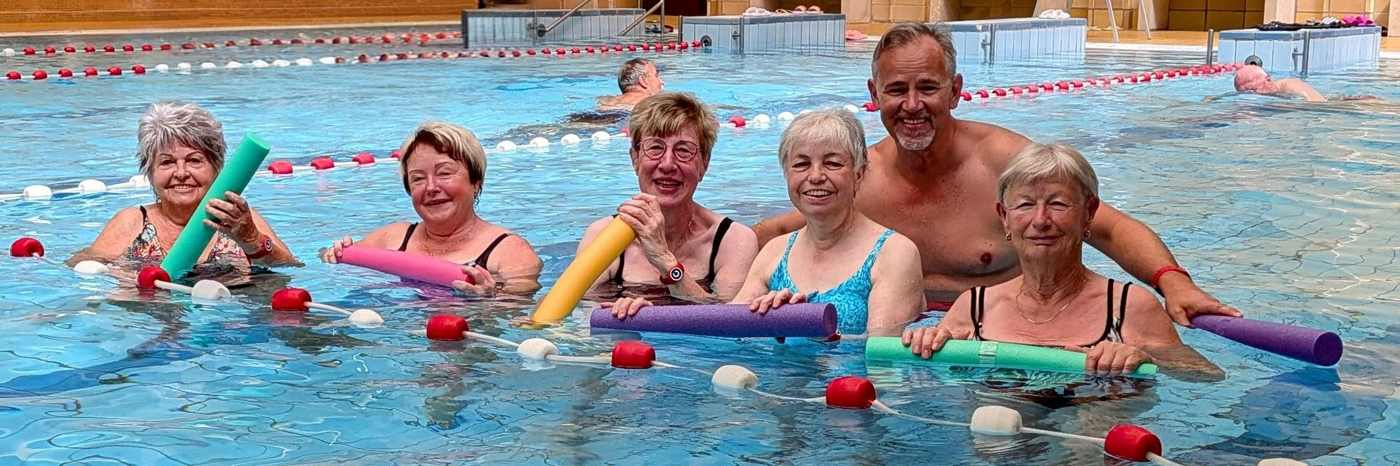 fünf Frauen und ein Mann stehen im Hallenbad im Wasser und lachen in die Kamera.