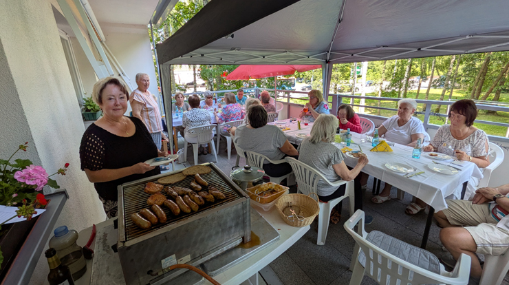 Viele ältere Menschen sitzen auf einer Terrasse unter Schirmen und Pavillons. Im Vordergrund steht ein Gasgrill mit Bratwürsten darauf.