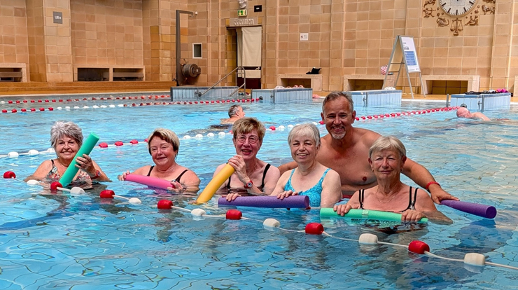 fünf Frauen und ein Mann stehen im Hallenbad im Wasser und lachen in die Kamera.
